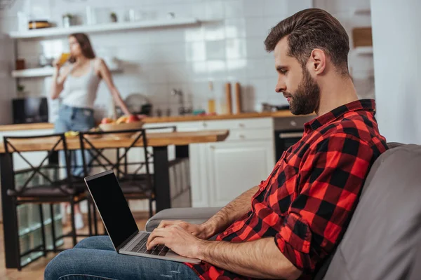 Enfoque selectivo de freelancer concentrado utilizando el ordenador portátil con pantalla en blanco en la cocina cerca de la novia en el fondo - foto de stock