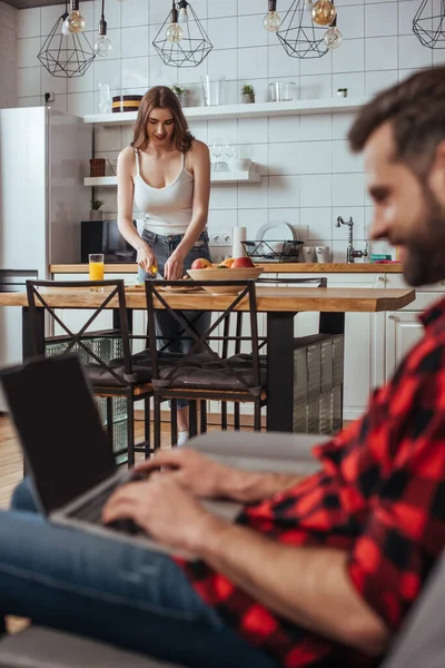 Enfoque selectivo de freelancer que trabaja en el ordenador portátil con pantalla en blanco en la cocina cerca de novia atractiva en el fondo - foto de stock