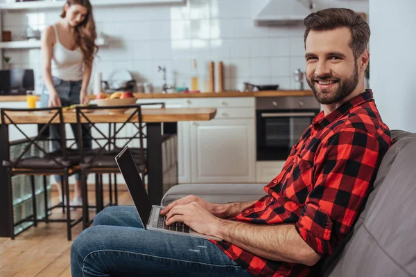 Messa a fuoco selettiva di bello freelance sorridente a macchina fotografica mentre lavora a computer portatile in cucina vicino a ragazza su sfondo — Foto stock