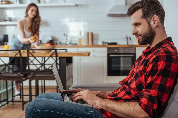 Enfoque selectivo de freelancer guapo que trabaja en el ordenador portátil en la cocina cerca de la novia en el fondo - foto de stock