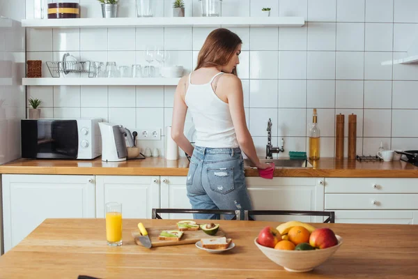 Vista trasera de la joven mujer preparando el desayuno en la cocina moderna cerca de la mesa con frutas frescas - foto de stock