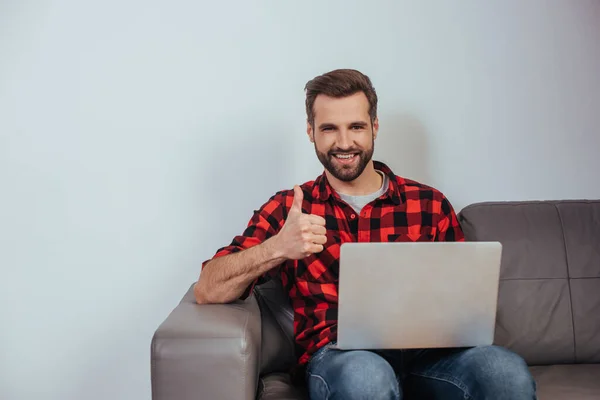 Feliz freelancer sonriendo a la cámara y mostrando el pulgar hacia arriba mientras está sentado en el sofá con el ordenador portátil - foto de stock