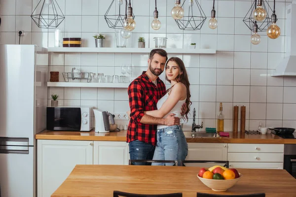 Счастливая молодая пара обнимается и смотрит в камеру, стоя на современной кухне — стоковое фото