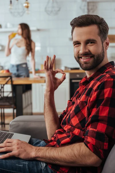 Foco seletivo de freelancer feliz olhando para a câmera e mostrando gesto ok perto da namorada em segundo plano — Fotografia de Stock