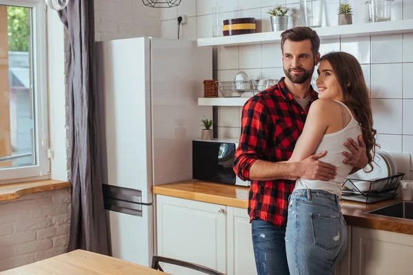 Felice giovane coppia che abbraccia e distoglie lo sguardo in cucina — Foto stock