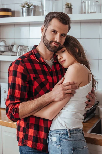 Hombre guapo sonriendo a la cámara mientras abraza atractiva novia en la cocina - foto de stock
