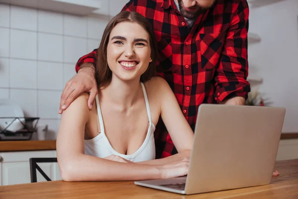 Обрезанный вид человека, трогающего плечо счастливой внештатной подружки, улыбающейся в камеру во время использования ноутбука — стоковое фото