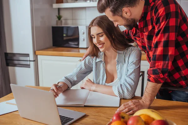 Junger Mann sitzt auf Tisch und berührt Schulter lächelnder Freundin, die mit Laptop und Notizbuch arbeitet — Stockfoto