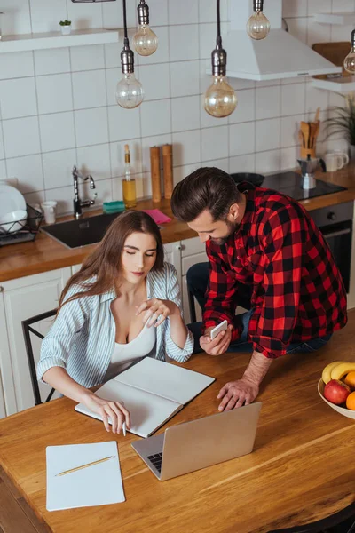 High angle view of man sitting on table and showing smartphone to serious girlfriend working on laptop — Stock Photo
