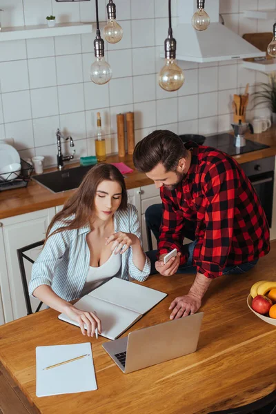 Vista de ángulo alto del hombre sentado en la mesa y mostrando el teléfono inteligente a la novia seria que trabaja en el ordenador portátil - foto de stock