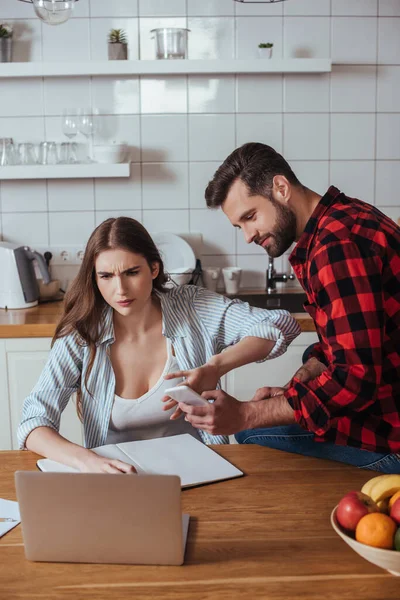 Smiling man sitting on table and showing smartphone to serious, busy girlfriend working on laptop — Stock Photo