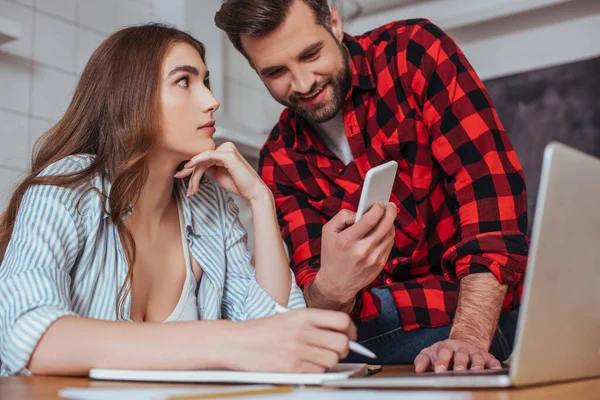 Sonriente hombre mostrando teléfono inteligente a novia seria trabajando en el ordenador portátil - foto de stock