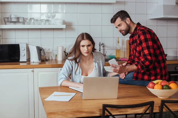 Hombre guapo sosteniendo teléfono inteligente mientras está sentado en la mesa cerca de novia seria trabajando en el ordenador portátil — Stock Photo