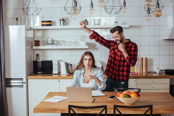 Fröhlicher Mann albert neben unzufriedener Freundin herum, die neben Laptop und Notizbuch in Küche sitzt — Stockfoto