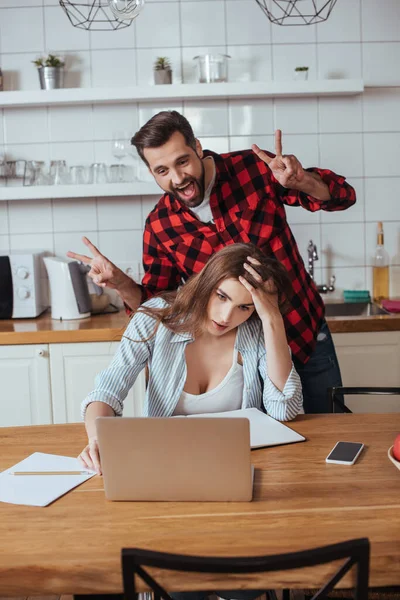 Веселый человек валяет дурака рядом с измученной девушкой, сидящей рядом с ноутбуком и ноутбуком на кухне — стоковое фото