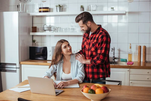 Hombre guapo tocando hombros de novia bonita sentada en la mesa cerca de la computadora portátil y portátil - foto de stock