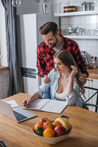 Schöner Mann berührt Schultern der attraktiven Freundin, schaut auf Laptop und schreibt in Notizbuch in der Küche — Stockfoto