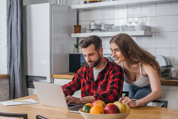 Selective focus of smiling girl standing near busy, serious boyfriend working on laptop — Stock Photo