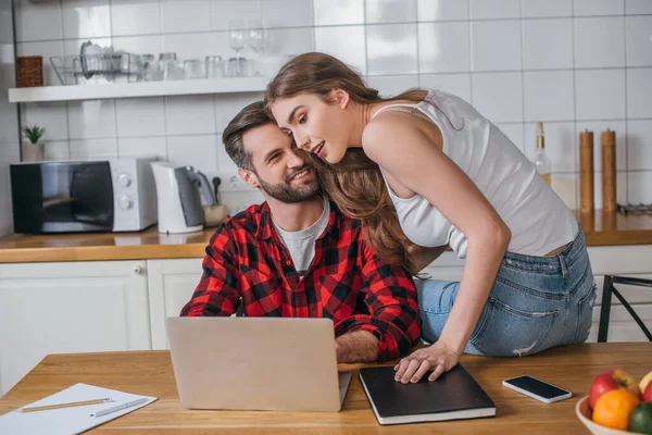 Belle fille assise sur la table près petit ami souriant travaillant sur ordinateur portable — Photo de stock