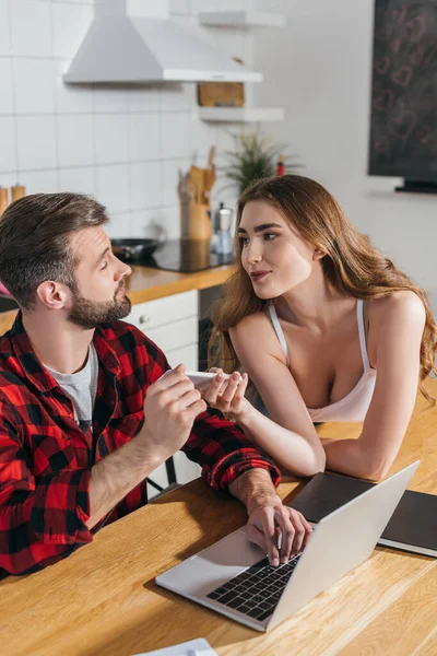 Belle fille souriante montrant smartphone occupé, petit ami sérieux assis à la table de cuisine et travaillant sur ordinateur portable — Photo de stock