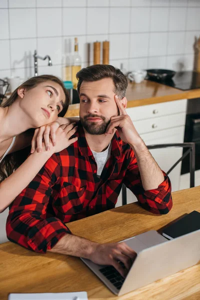 Bored girl leaning on shoulder of busy boyfriend sitting at kitchen table and working on laptop — Stock Photo