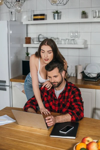 Красивая девушка опирается на плечо занятого парня, сидящего за кухонным столом и работающего на ноутбуке — стоковое фото