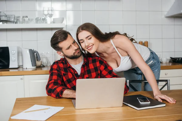 Cheerful girl looking at laptop screen near freelancer working while sitting at kitchen table — Stock Photo