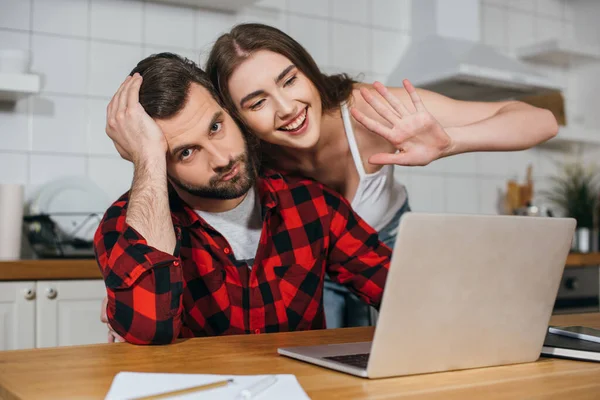 Serious freelancer looking at camera and touching head while cheerful girlfriend waving hand at laptop screen — Stock Photo