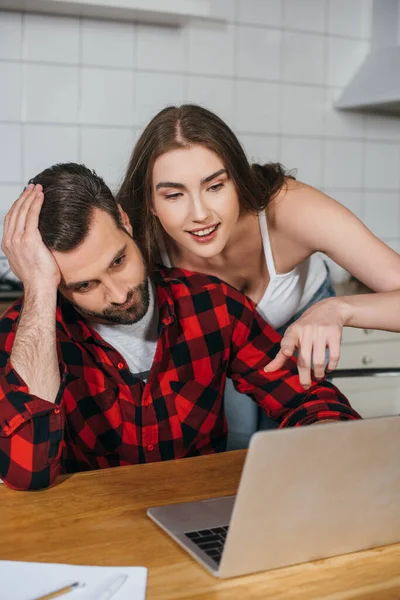 Smiling girl point with finger at laptop screen near tired boyfriend — Stock Photo