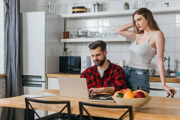 Привлекательная девушка, стоящая рядом с парнем сидя за кухонным столом и работая на ноутбуке — стоковое фото