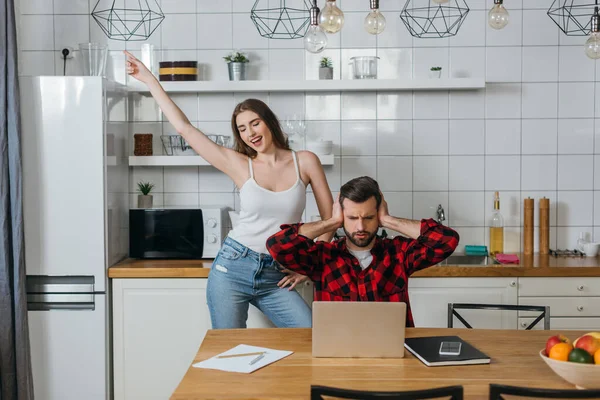 Cheerful girl fooling around and dancings near boyfriend covering ears with hands while sitting near laptop and papers — Stock Photo