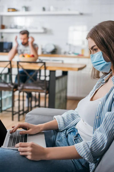 Foco seletivo de jovem freelancer em máscara médica trabalhando no laptop na cozinha perto do namorado em segundo plano — Fotografia de Stock
