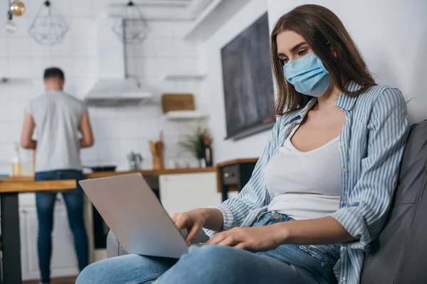 Foco seletivo de jovem freelancer em máscara médica trabalhando no laptop na cozinha perto do namorado em segundo plano — Fotografia de Stock