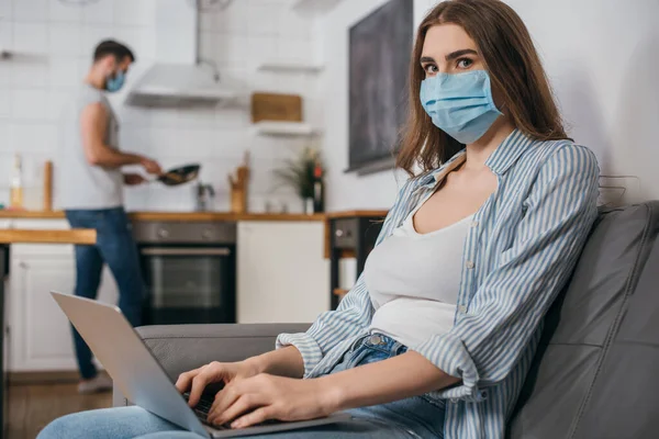 Selektiver Fokus der Freiberuflerin in medizinischer Maske, die am Laptop in der Küche arbeitet, in der Nähe ihres Freundes im Hintergrund — Stockfoto