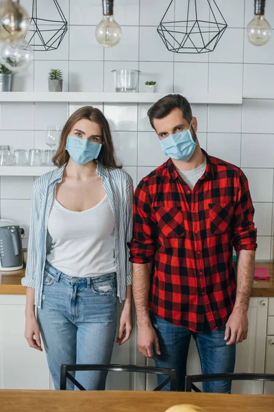 Молода пара в медичних масках дивиться на камеру, стоячи на сучасній кухні — стокове фото