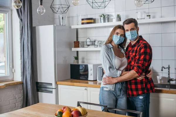 Jovem casal em máscaras médicas abraçando e olhando para a câmera na cozinha moderna — Fotografia de Stock