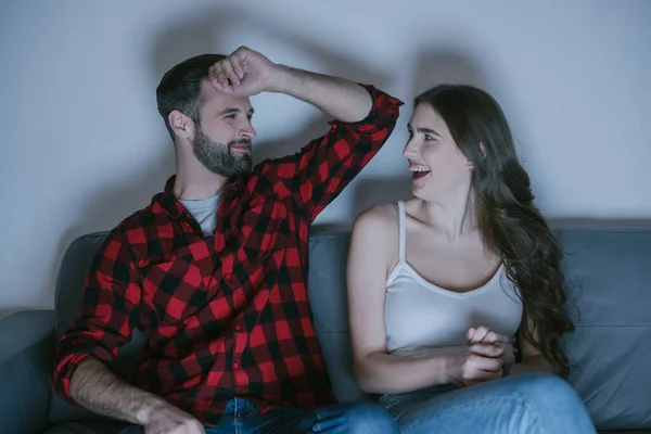 Casal feliz olhando um para o outro e rindo enquanto assiste tv no sofá em casa — Fotografia de Stock