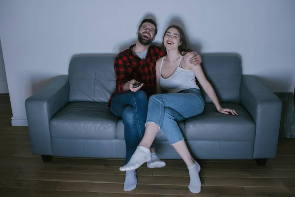 Веселая пара смеется, смотря телевизор на диване дома — стоковое фото