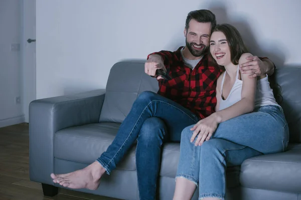 Alegre pareja riendo y el hombre señalando con el dedo mientras mira la televisión en el sofá en casa - foto de stock