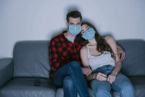Jeune couple dans des masques médicaux regarder la télévision sur le canapé à la maison — Photo de stock