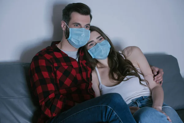 Молодая пара в медицинских масках смотрит телевизор на диване дома — стоковое фото