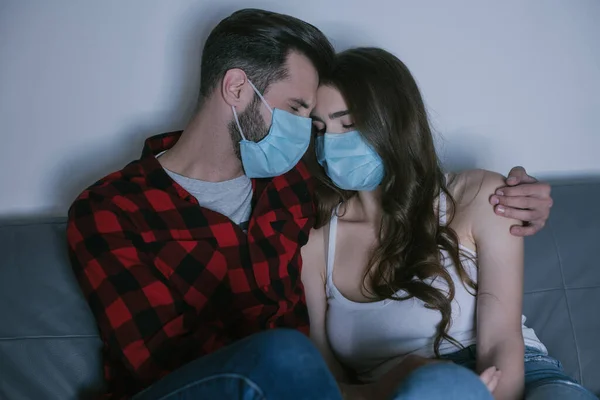 Couple déprimé dans des masques médicaux assis avec les yeux fermés tout en regardant la télévision — Photo de stock