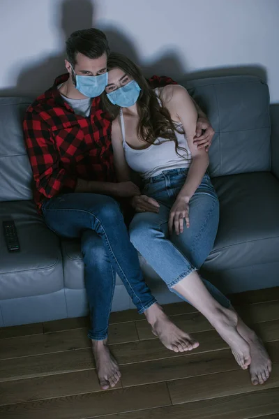 Vue grand angle de jeune couple dans des masques médicaux regarder la télévision — Photo de stock