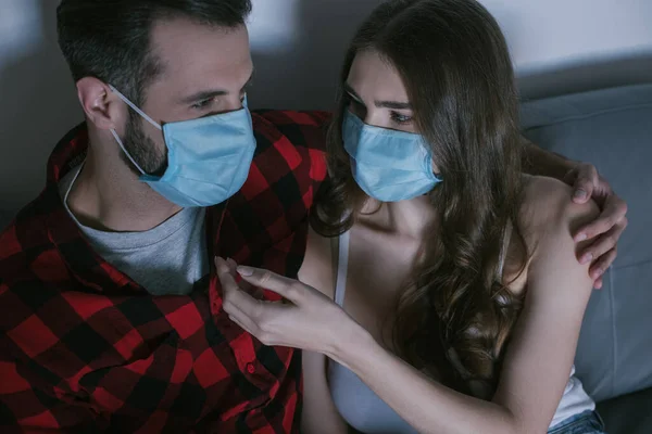 Jeune couple en masques médicaux se regardant tout en regardant la télévision — Photo de stock