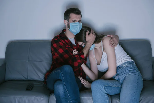Joven pareja en médico máscaras viendo tv mientras hombre calmando molesto chica - foto de stock