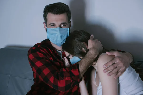 Молодая пара в медицинских масках смотрит телевизор, пока мужчина обнимает расстроенную девушку — стоковое фото