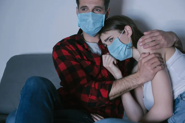 Joven pareja en médico máscaras viendo tv mientras hombre abrazando triste chica - foto de stock