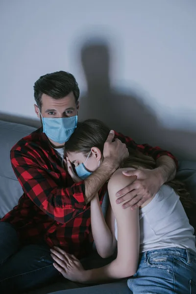 Joven pareja en médico máscaras viendo tv mientras hombre abrazando molesto chica - foto de stock