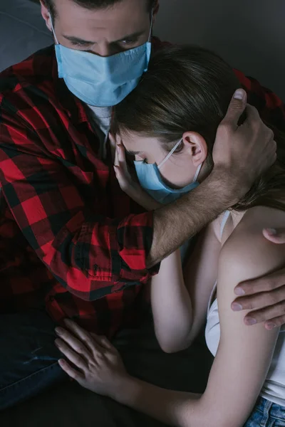 Young couple in medical masks watching tv while man embracing upset girlfriend — Stock Photo