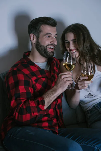 Glückliches junges Paar lacht, während es auf dem Sofa sitzt und Weißweingläser hält — Stockfoto